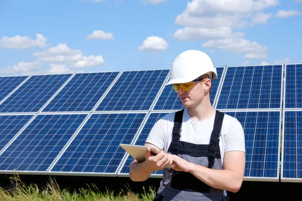 Solar panels engineer in white cask, and grey outfit stadnding near solar panels field and typing on tablet. Hot sunny summer weather. Copy space. Concept of renewable and clean energy, electricity.