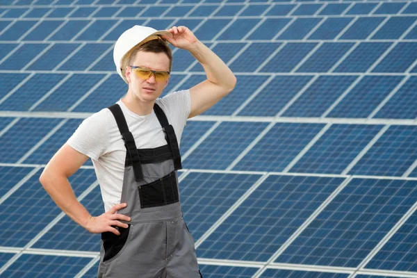 Electrical worker holding white safety hat and standing at power plant. Solar engineer in protective yellow glasses and grey overalls stadnding near solar panels field. Concept of renewable and clean energy, electricity, technology. People at work.