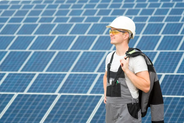 Solar panels engineer in white cask, protective yellow glasses and grey outfit stadnding near solar panels field. Hot sunny summer weather. Alternative types of energy. Concept of renewable and clean energy, technology. Man at work