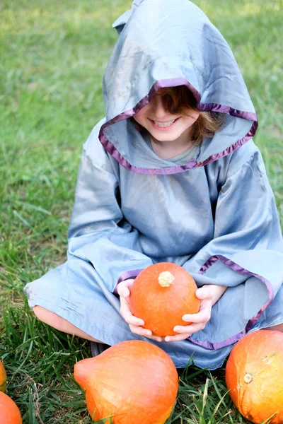 Boy in wizard costume sits on the lawn, holds pumpkin and smiles. Halloween concept.