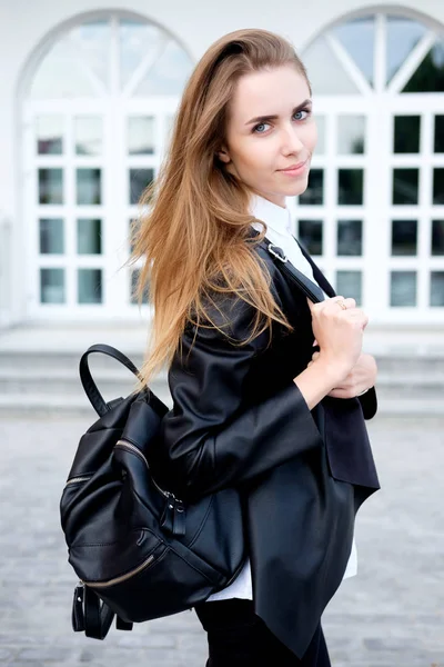 Portrait of stylish beautiful young woman in fashionable black leather jacket, white shirt, black trousers holding black backpack.