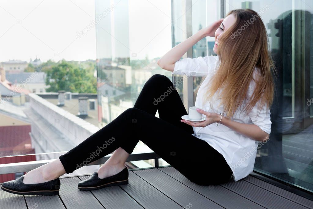 Portrait of young funny cool girl or model in stylish white shirt and black trousers holding cup of coffee and laughing  in the open air place or observation deck. Old town on background. Concept love morning.