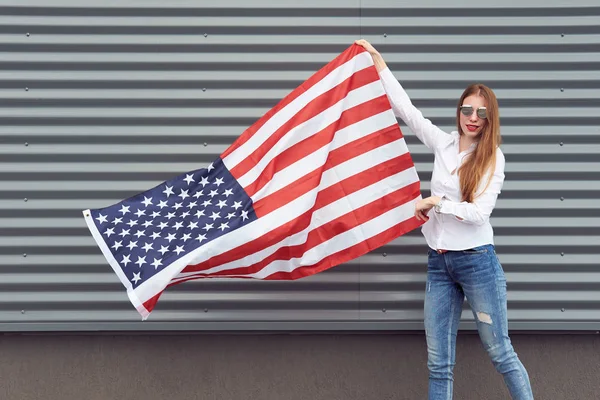 White redhead woman with red painted lips holding waved by wind usa flag. Grey metal panel background. Independence day and patriotic concept.