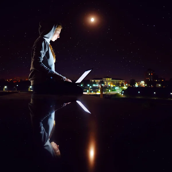 Young white redhead skinny girl, weared in grey hoody, sitting outdoors and working with laptop in night, cityscape background.