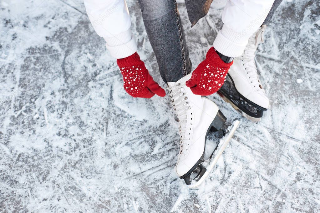 Girl tying shoelaces on ice skates before skating on the ice rink, hands in red knitted gloves. View from top.