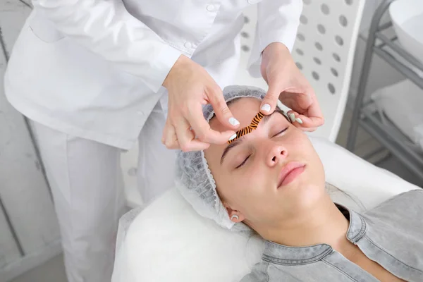 Cosmetologist making taping face procedure using tiger colored tapes in beauty parlor. Young attractive woman or model with closed eyes lying on the procedure table. Concept beauty.