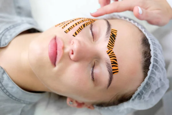 Cosmetologist making taping face procedure using tiger colored tapes in beauty parlor. Young attractive woman or model with closed eyes lying on the procedure table. Concept beauty.