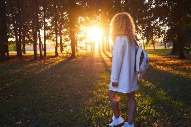 Kızıl saçlı kıvırcık zencefil kız, beyaz sırt çantası ile beyaz giysili renksizdir park akşam ve günbatımı ışığı izlerken bir yürüyüşü vardır. Arkadan görüntülemek.