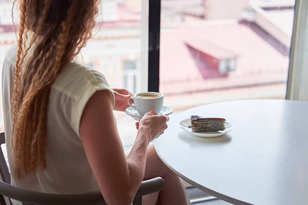 Curly redhead skinny woman sitting in cafe in front of window with cup of coffee and observing european cityscape