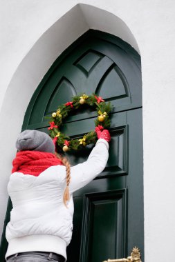 kız kırmızı eşarp ve beyaz ceket Noel çelenk yeşil kapıda asılı. Tatil.