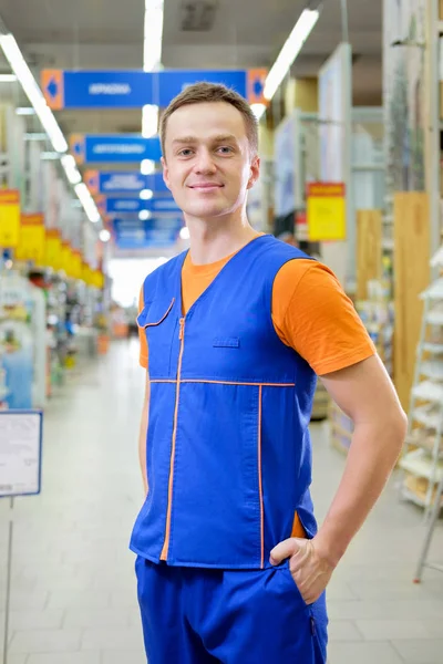 Young salesman standing at construction super store. Portrait of attractive confident working man in uniform on his workplace in building store.