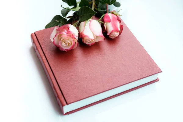 Three beautiful roses lies on red leather covered wedding album or wedding book. Wedding book lie on white background. Top view.