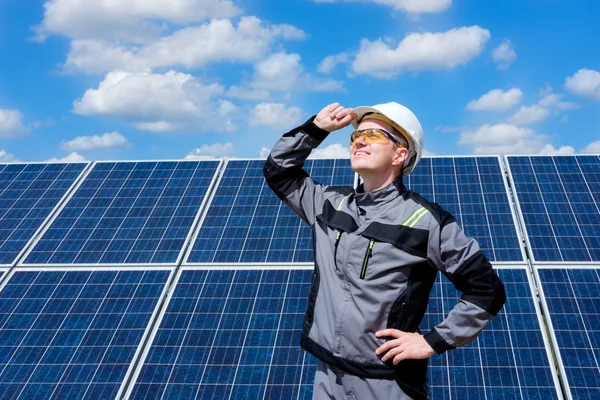 Solar panels engineer in white cask, protective yellow glasses and grey outfit standing near solar panels field and watching at the sun. Hot sunny summer weather. Solar panels field background. Concept of renewable and clean energy, electricity, tech