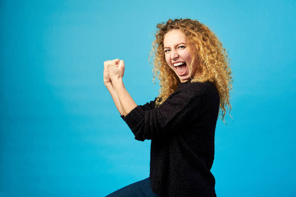 Elated satisfied young redhead curly woman celebrating and cheering a success while punching the air with her fists over blue wall background. Yes. I did it. Positive feelings. Studio shot.