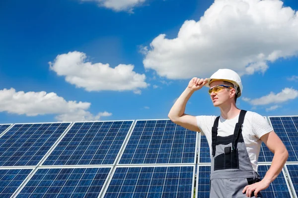 Solar panels engineer in white cask, protective yellow glasses and grey outfit standing near solar panels field and watching at the sun. Hot sunny summer weather. Solar panels field and sky background. Concept of renewable and clean energy, electrici