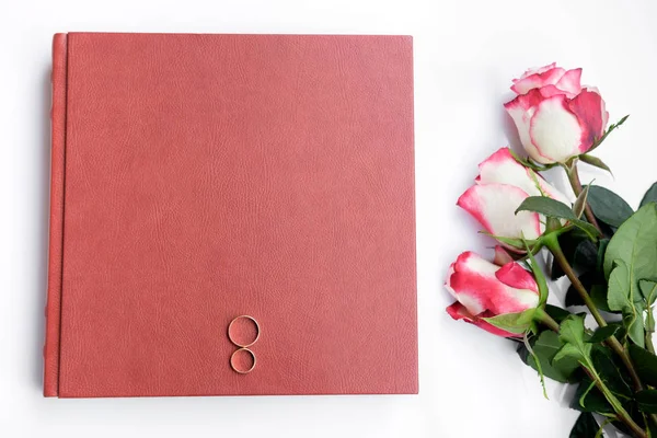 Red leather covered wedding book or album with two wedding rings and three roses lies on white background. Wedding memories. Love symbols. Top view.