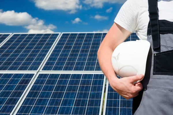 Solar panels engineer wearing grey outfit holding white protective cask, hand close, solar pannel field and sky background. Hot sunny summer weather. Copy space. Concept of renewable and clean energy, technology.