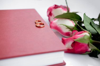 Düğün kitap kırmızı deri kaplı ya da albüm ile iki Alyans beyaz arka plan üzerinde yatıyor. Güzel üç gül buket düğün kitabı yatıyor. Düğün anıları. Aşk sembolleri. Üstten görünüm. Yakın çekim.