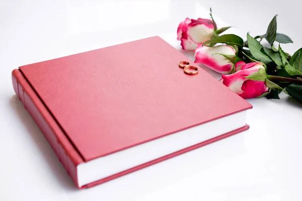 Red leather covered wedding book or album with two wedding rings lies on white background. Beautiful bouquet of three roses lies near wedding book. Wedding memories. Symbols of love. Top view. Close up.