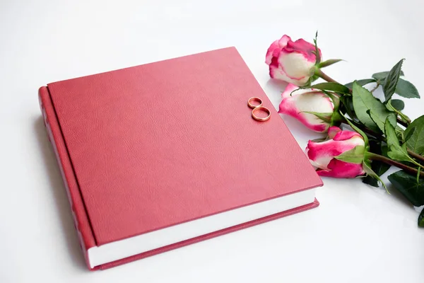 Red leather covered wedding book or album with two wedding rings lies on white background. Beautiful bouquet of three roses lies near wedding book. Wedding memories. Symbols of love. Top view. Close up.