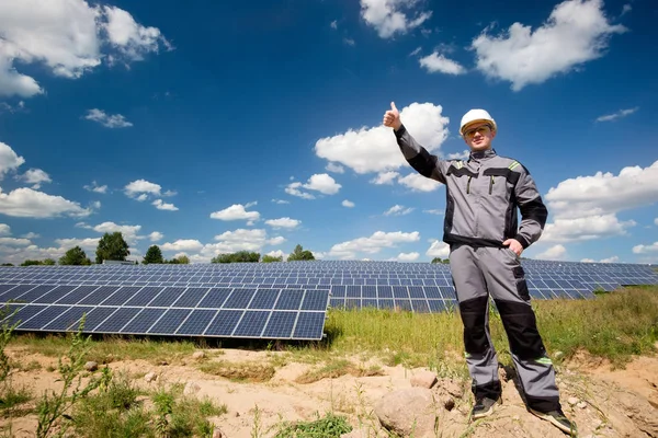 Solar panels engineer or electrician in white cask, protective yellow glasses and grey outfit standing near solar panels field and showing thumbs up. Solar panels field and sky background. Concept of renewable and clean energy, electricity, technolog