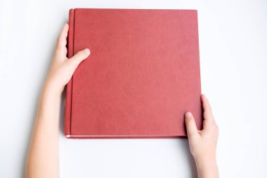Kırmızı deri kaplı photobook veya albüm tutan çocuk. Photobook beyaz parlak zemin üzerine yalan. Üstten görünüm.