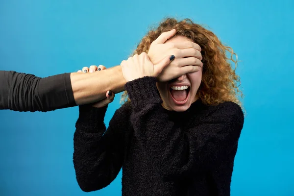 Man\'s hand closing eyes of young redhead curly woman. Woman screaming and trying to break free from man\'s hand. Studio shot on blue background. Concept of violence and cruelty in relationships.