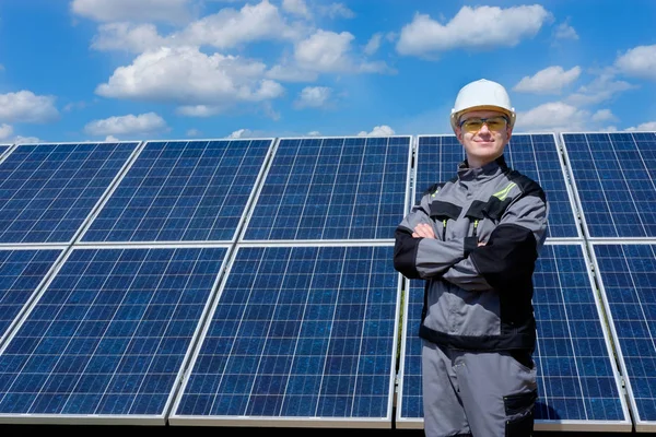 Solar panels engineer in white cask, protective yellow glasses and grey outfit standing near solar panels field. Solar panels field and sky background. Concept of renewable and clean energy, electricity, technology. Worker at workplace.