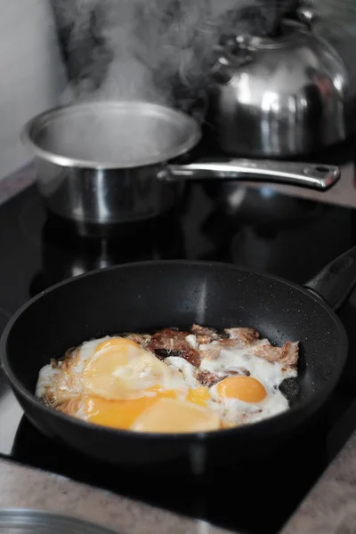 Los huevos fritos se cocinan en una sartén, el ganado está hirviendo agua en la parte posterior —  Fotos de Stock