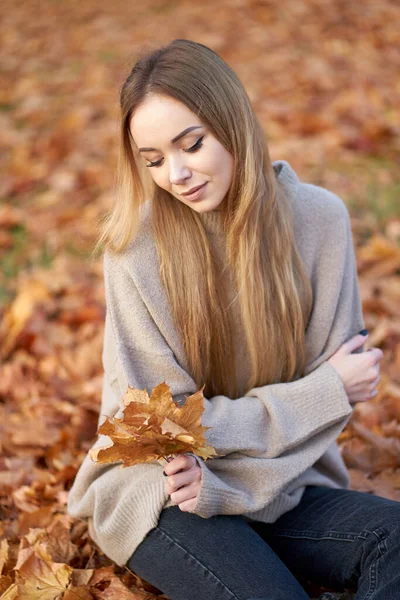 美しい長い髪をしたブロンドの若い女の子は 流行の居心地の良いセーターと黒いジーンズを手にメープルの葉の花束を持っています 秋の時間だ 肖像画 — ストック写真