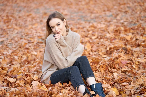 秋は毎日探します 美しいですスタイリッシュなブロンド自信のある女の子身に着けているセーター 黒ジーンズと革黒ブーツで秋の公園に座って — ストック写真
