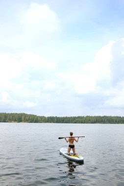 Siyah mayo giyen genç bir kadın SUP tahtasının üzerinde gölde yüzüyor.. 