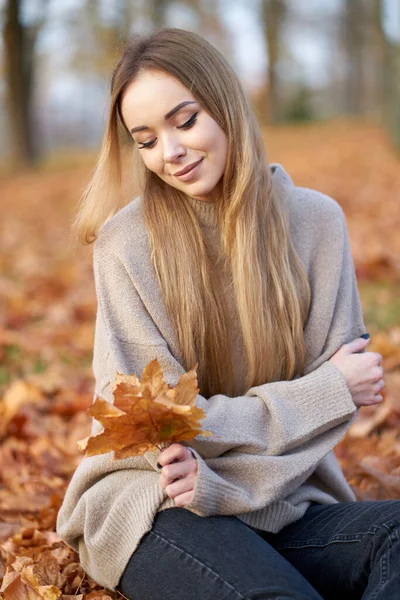 魅力的な笑顔と長いブロンドの美しい髪を持つスタイリッシュな魅力的な若い女性手の中にオレンジメープルの葉を保持流行の自動セーターを着て — ストック写真