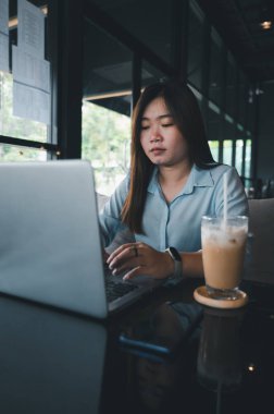 Tayland kökenli bir Asyalı kadın ofis çalışanı gün içinde bir kahve dükkanında not defterleriyle yazıp çalışıyor. Mavi bir gömlek giyiyordu.