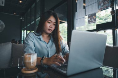 Tayland kökenli bir Asyalı kadın ofis çalışanı gün içinde bir kahve dükkanında not defterleriyle yazıp çalışıyor. Mavi bir gömlek giyiyordu.