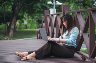 Parktaki tahta köprüde dizüstü bilgisayar kullanan kahverengi saçlı bir kadının yan görüntüsü. Genç bir ofis çalışanı gün batımında parkta yeşil çimlerin üzerinde telefonla konuşuyor..