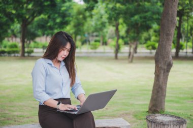 Parkta dizüstü bilgisayar ya da bilgisayar kullanan Asyalı bir ofis çalışanı. Mavi bir gömlek giyiyor..