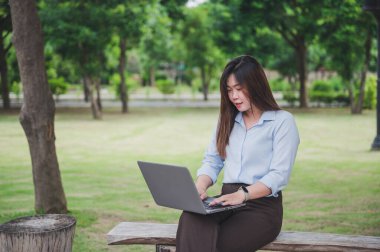 Parkta dizüstü bilgisayar ya da bilgisayar kullanan Asyalı bir ofis çalışanı. Mavi bir gömlek giyiyor..