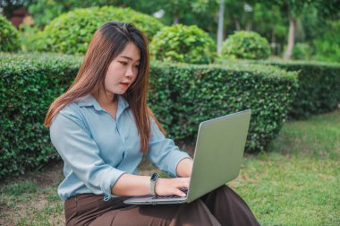 Parkta dizüstü bilgisayar ya da bilgisayar kullanan Asyalı bir ofis çalışanı. Mavi bir gömlek giyiyor..