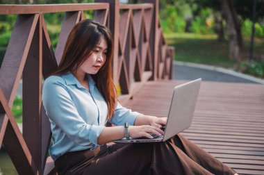 Parktaki tahta köprüde dizüstü bilgisayar kullanan kahverengi saçlı bir kadının yan görüntüsü. Genç bir ofis çalışanı gün batımında parkta yeşil çimlerin üzerinde telefonla konuşuyor..