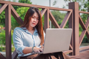 Parktaki tahta köprüde dizüstü bilgisayar kullanan kahverengi saçlı bir kadının yan görüntüsü. Genç bir ofis çalışanı gün batımında parkta yeşil çimlerin üzerinde telefonla konuşuyor..