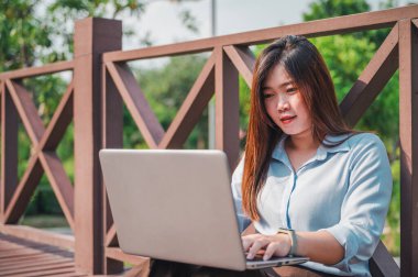Parktaki tahta köprüde dizüstü bilgisayar kullanan kahverengi saçlı bir kadının yan görüntüsü. Genç bir ofis çalışanı gün batımında parkta yeşil çimlerin üzerinde telefonla konuşuyor..