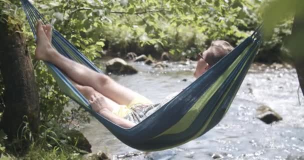 Man kopplar av i en Hammock nära floden. Sakta i backarna. Stilig man dagdrömmar, landsbygd natur med skog i bakgrunden. — Stockvideo