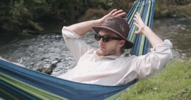 特写视图。夏天的一天，戴着帽子和太阳镜的英俊男子躺在吊床上摇晃着 — 图库视频影像