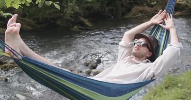 夏天的一天，戴着帽子和太阳镜的英俊男子躺在吊床上摇晃着 — 图库视频影像