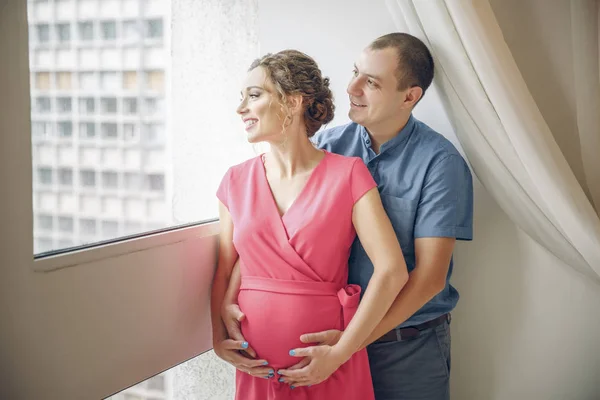 Pregnancy and people concept - happy man hugging his pregnant wife standing at window — Stock Photo, Image