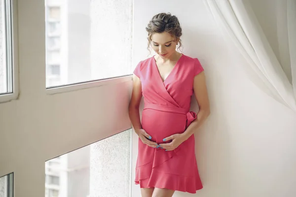 young pregnant woman staying near the window in pink dress. in anticipation of the child