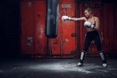 Kadın boksör boks stüdyosunda büyük bir kum torbasına vuruyor. Kadın boksör sıkı çalışıyor..