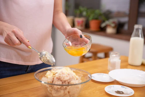 Stages of preparation of meat glomeruli. The woman adds a chicken egg to the stuffing. Next to the table is a dough and tools.