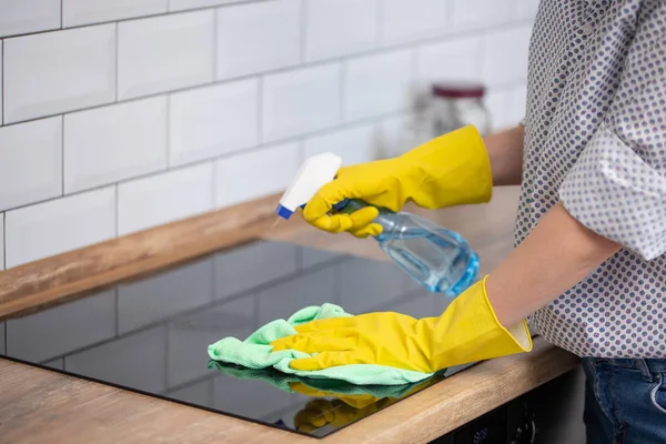 Close up van menselijke hand met beschermende handschoenen inductie kookplaat met doek schoonmaken en houden sproeiflacon. — Stockfoto